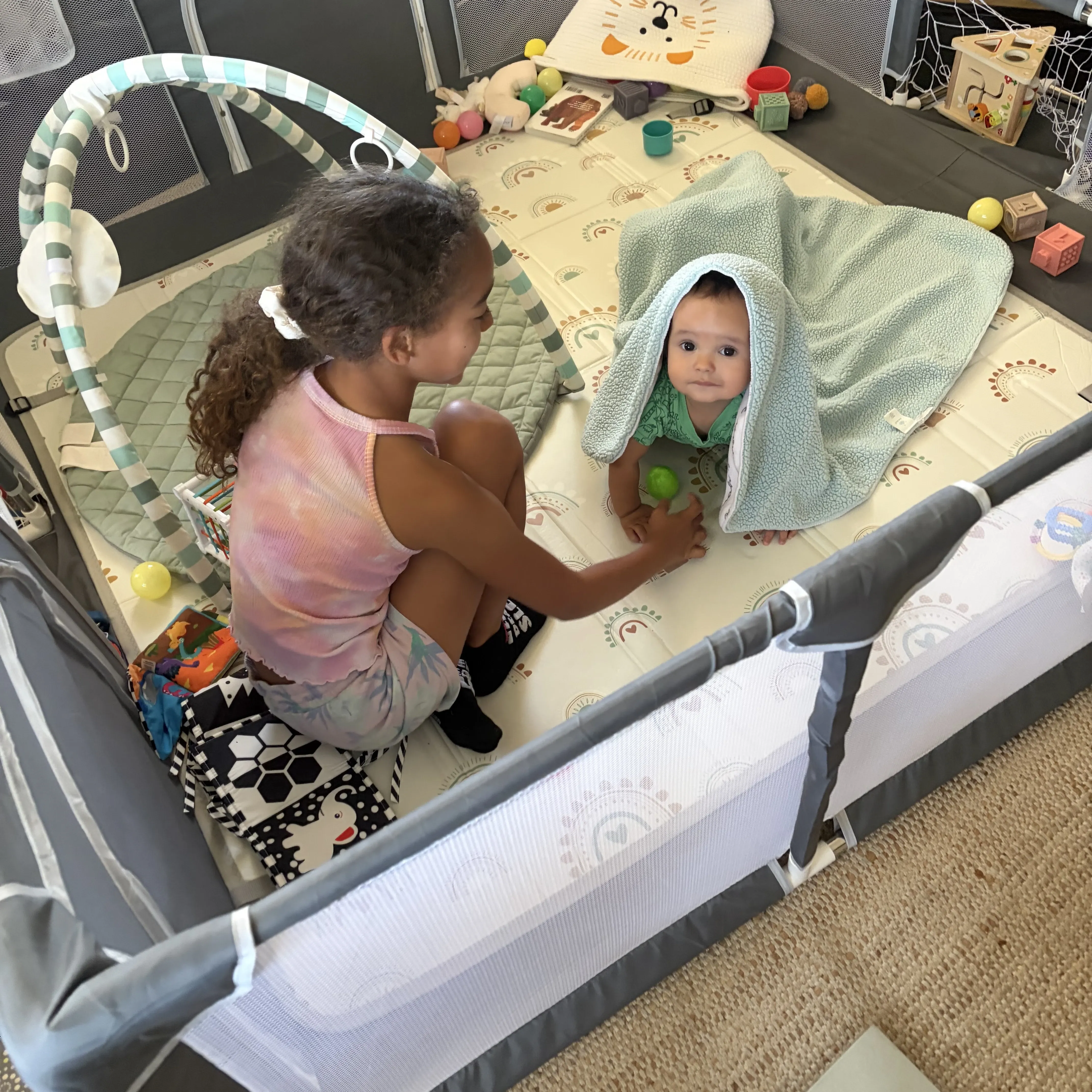 Niabelle sitting with Noah in the playpen, handing him a ball while he peeks out from under a blanket